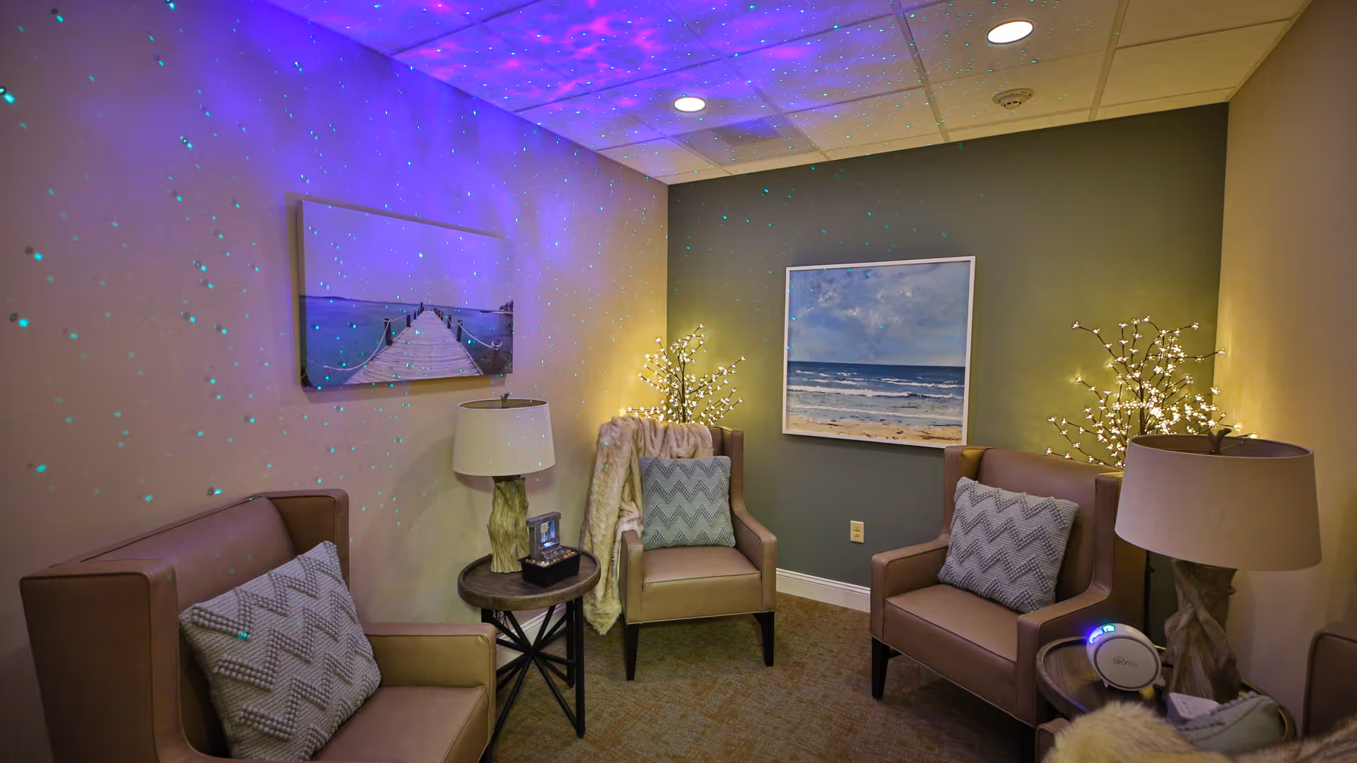 A cozy sitting room with three beige armchairs, each with a patterned cushion. Two small round side tables hold decorative lamps and a small device. The walls are decorated with two framed pictures, one of a beach scene and the other of a wooden pier. Soft lighting and star-like projections create a calming atmosphere.