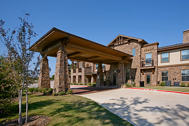 Exterior view of a senior living facility building with stone and beige walls, featuring a covered entrance supported by stone pillars, a driveway, and landscaped grass and trees under a clear blue sky.