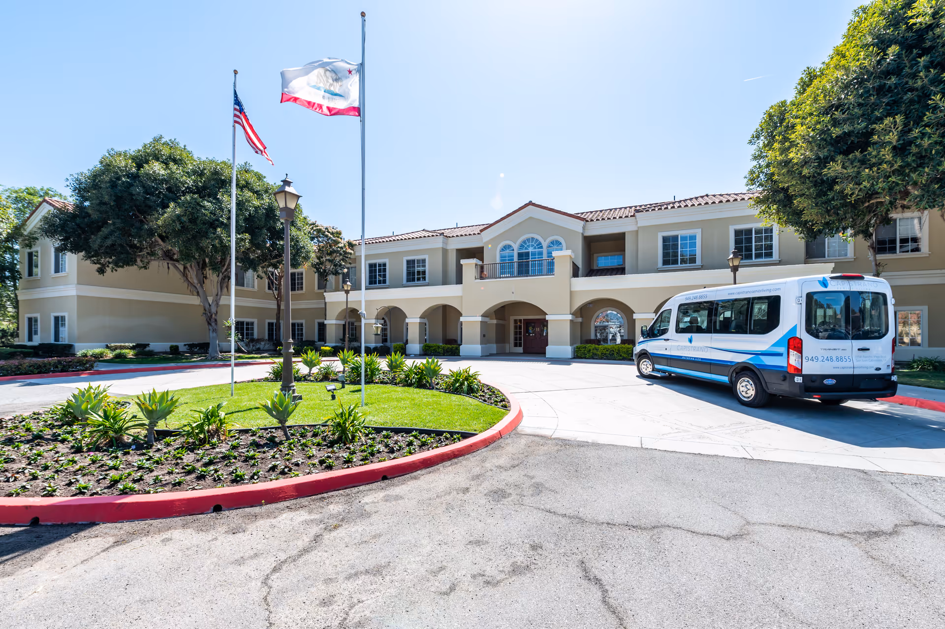 Exterior view of San Juan Hills Healthcare Center, a two-story building with beige walls and a red-tiled roof. There are two flagpoles with the American and California state flags, a landscaped circular driveway with plants, and a white shuttle van parked near the entrance under a clear blue sky.