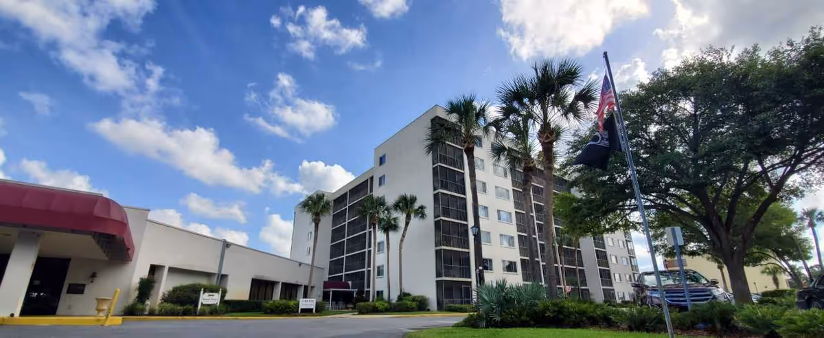 Wide-angle view of Woodland Towers' multi-story building front with a covered entrance, palm trees, flagpoles, and a blue sky.