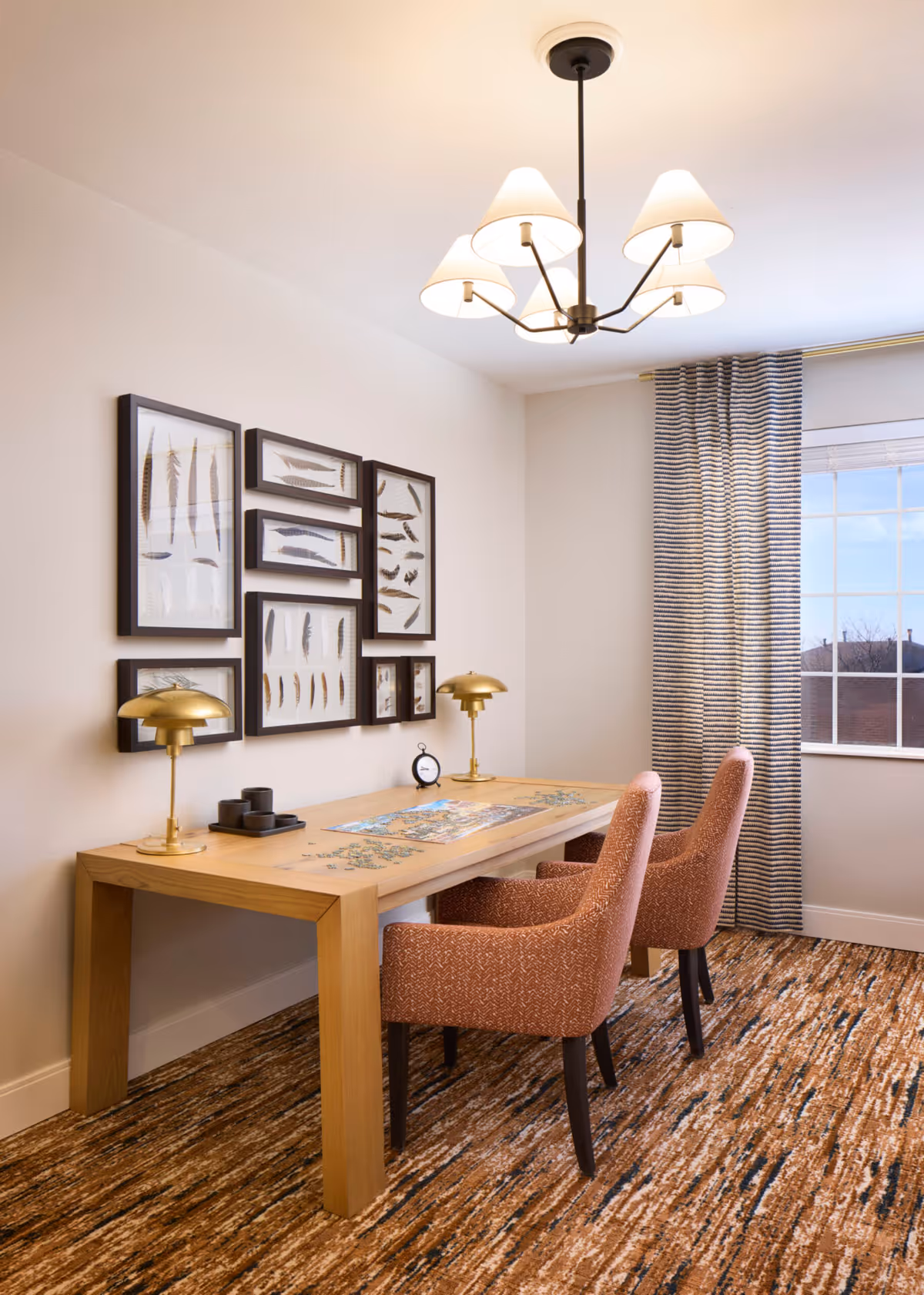 Bright interior room with a wooden table, two upholstered chairs, brass desk lamps, framed feather artwork on the wall, and a window with striped curtains.