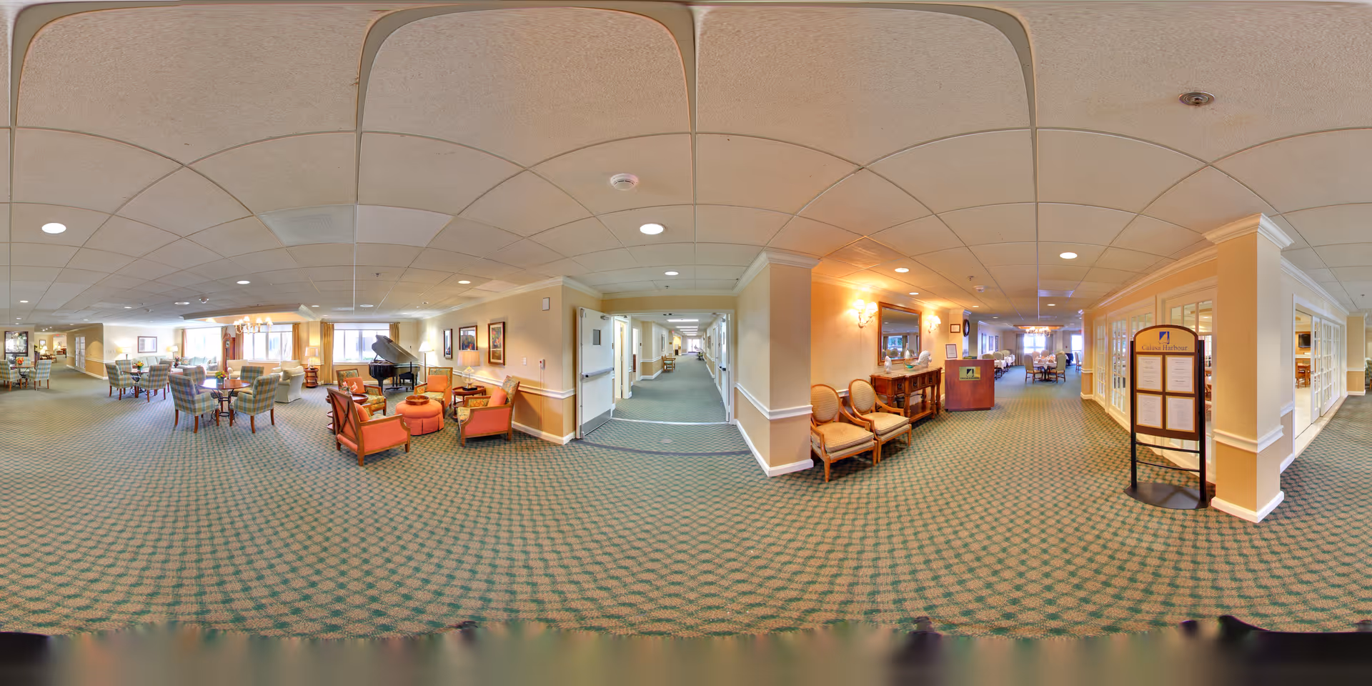 A spacious and well-lit senior living facility common area with patterned green carpet and beige walls. The room features multiple seating arrangements including armchairs and small tables, a grand piano, framed artwork on the walls, and a hallway leading further into the facility. There is a wooden console table with chairs and a standing signboard displaying information near the entrance to another room.