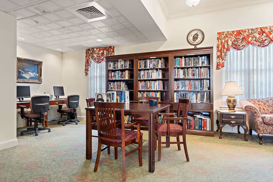 A cozy library and computer area in a senior living facility with a wooden table and four chairs in the center, a large bookshelf filled with books against the wall, two computer desks with chairs and monitors on the left, floral curtains on the windows, a side table with a lamp, and a patterned sofa on the right.