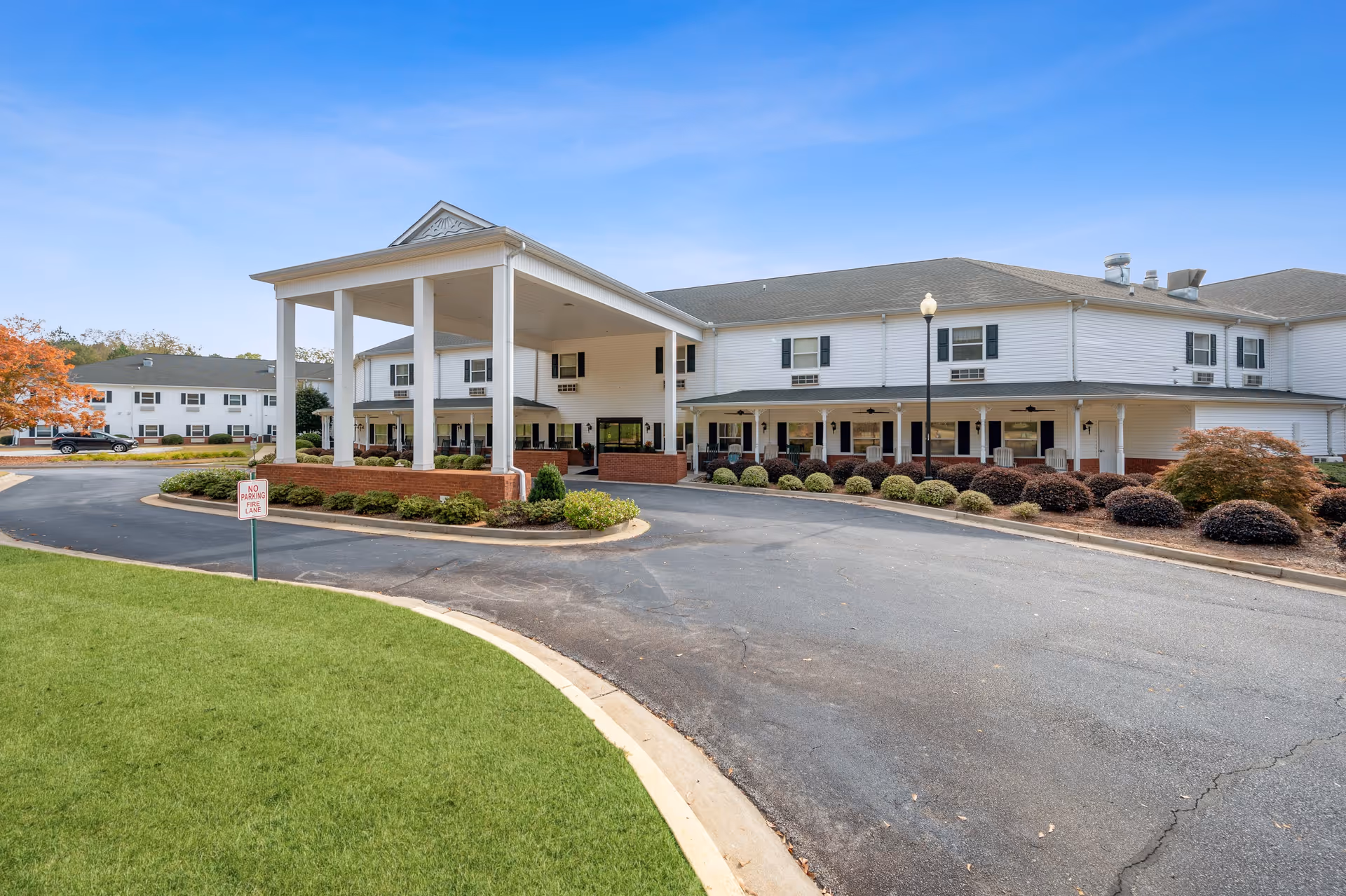 Exterior view of a two-story senior living facility with white siding and a covered entrance supported by tall white columns. The driveway curves around landscaped areas with bushes and a green lawn under a clear blue sky.