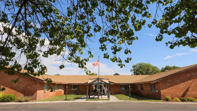 Front exterior view of a single-story brick building with a central entrance covered by a small canopy. An American flag is flying on a flagpole in front of the entrance. The building is surrounded by green grass and some bushes, with tree branches framing the top of the image against a blue sky with a few clouds.