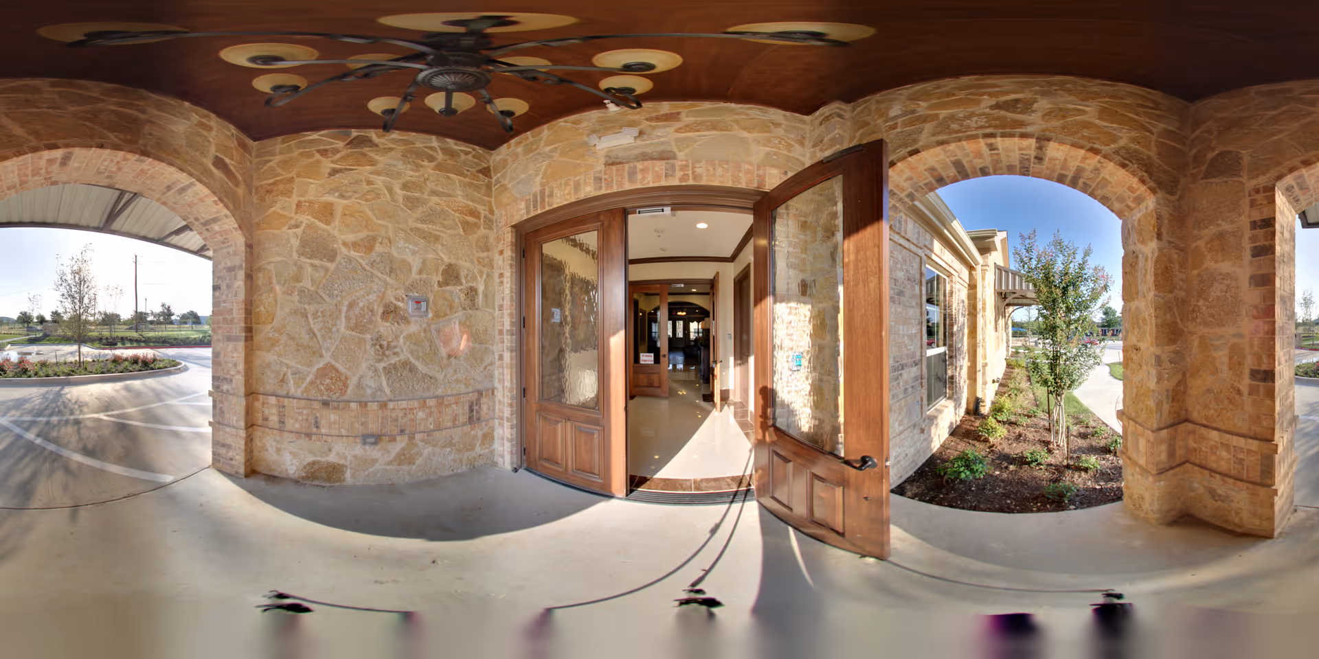 Stone exterior entrance of Avalon Memory Care facility with an open wooden door leading inside. The area features stone archways, a ceiling fan, and landscaped garden beds with small trees and shrubs. Sunlight casts shadows on the concrete floor and walls.