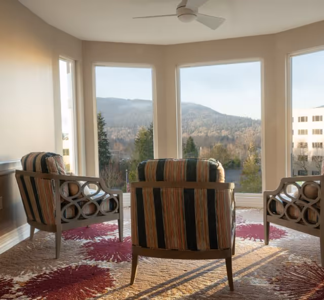 A cozy sitting area with three striped armchairs arranged in a semi-circle facing large windows that offer a scenic view of forested hills and a building in the distance. The room has a ceiling fan and a carpet with a floral pattern in shades of red and beige.