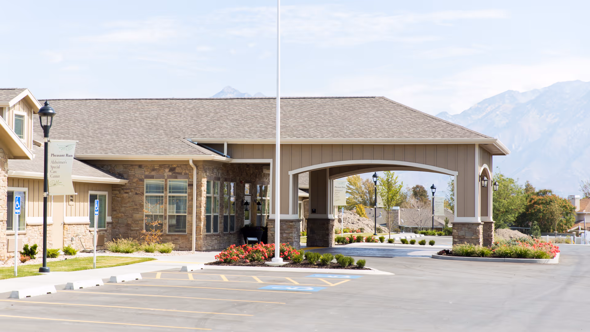 Exterior view of a single-story assisted living facility building with stone and beige siding, a covered entrance, landscaped flower beds, and a parking lot with handicap spaces. Mountains are visible in the background under a partly cloudy sky.