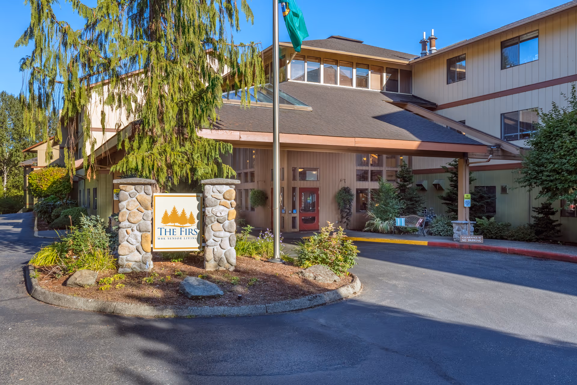 Front entrance of The Firs senior living facility with a stone sign, flagpole, driveway, and landscaped planting beds.