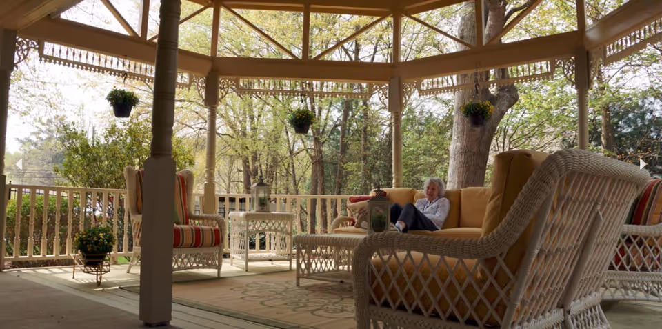 A covered outdoor patio area with white wicker furniture including chairs and a sofa with yellow cushions. A woman is sitting on the sofa. The patio is surrounded by trees and hanging flower pots.