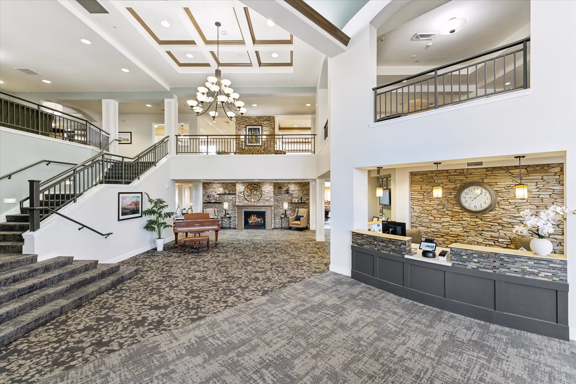 Spacious and well-lit senior living facility lobby with a patterned carpet, a staircase with black railings, a grand piano, a stone fireplace with a clock above it, and a reception desk with a stone accent wall and a large clock. The ceiling features recessed lighting and a chandelier.