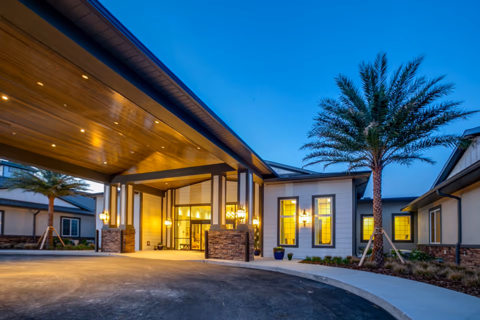 Exterior view of a senior living facility entrance at dusk with warm lights illuminating the covered driveway and building facade, featuring palm trees and a clear blue sky.