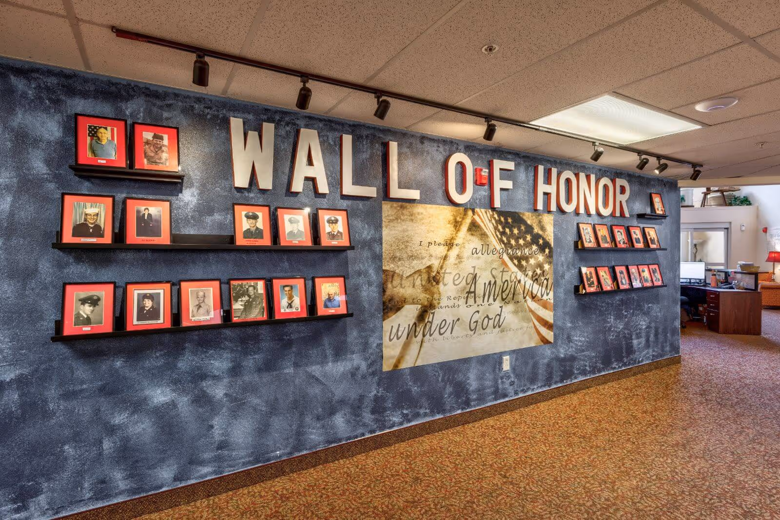 Interior view of a hallway featuring a 'Wall of Honor' display with framed photographs of veterans on shelves against a textured blue wall. The wall also has a large image of an American flag with the words 'I pledge allegiance to the United States of America under God.' Track lighting illuminates the display, and an office area is visible in the background.