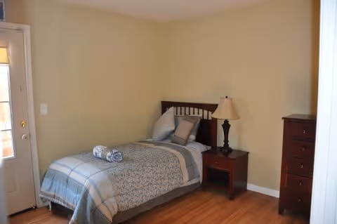A small bedroom with a single bed made with a patterned comforter and pillows. There is a rolled towel placed on the bed. Next to the bed is a wooden nightstand with a lamp. A tall wooden dresser is positioned against the wall. The room has light-colored walls and wooden flooring. A door with glass panels is visible on the left side.