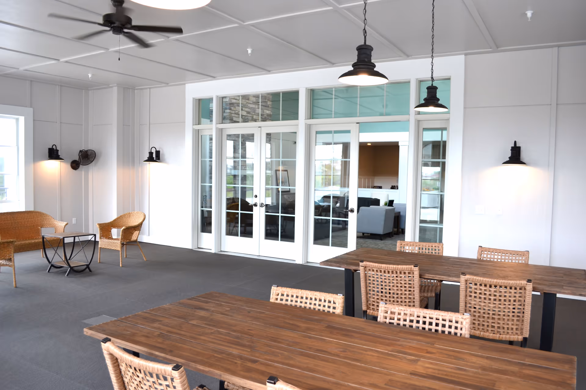 A spacious indoor seating area with wooden tables and wicker chairs. The room features white walls, ceiling fans, and hanging pendant lights. There are glass double doors leading to another room with additional seating visible through the glass.