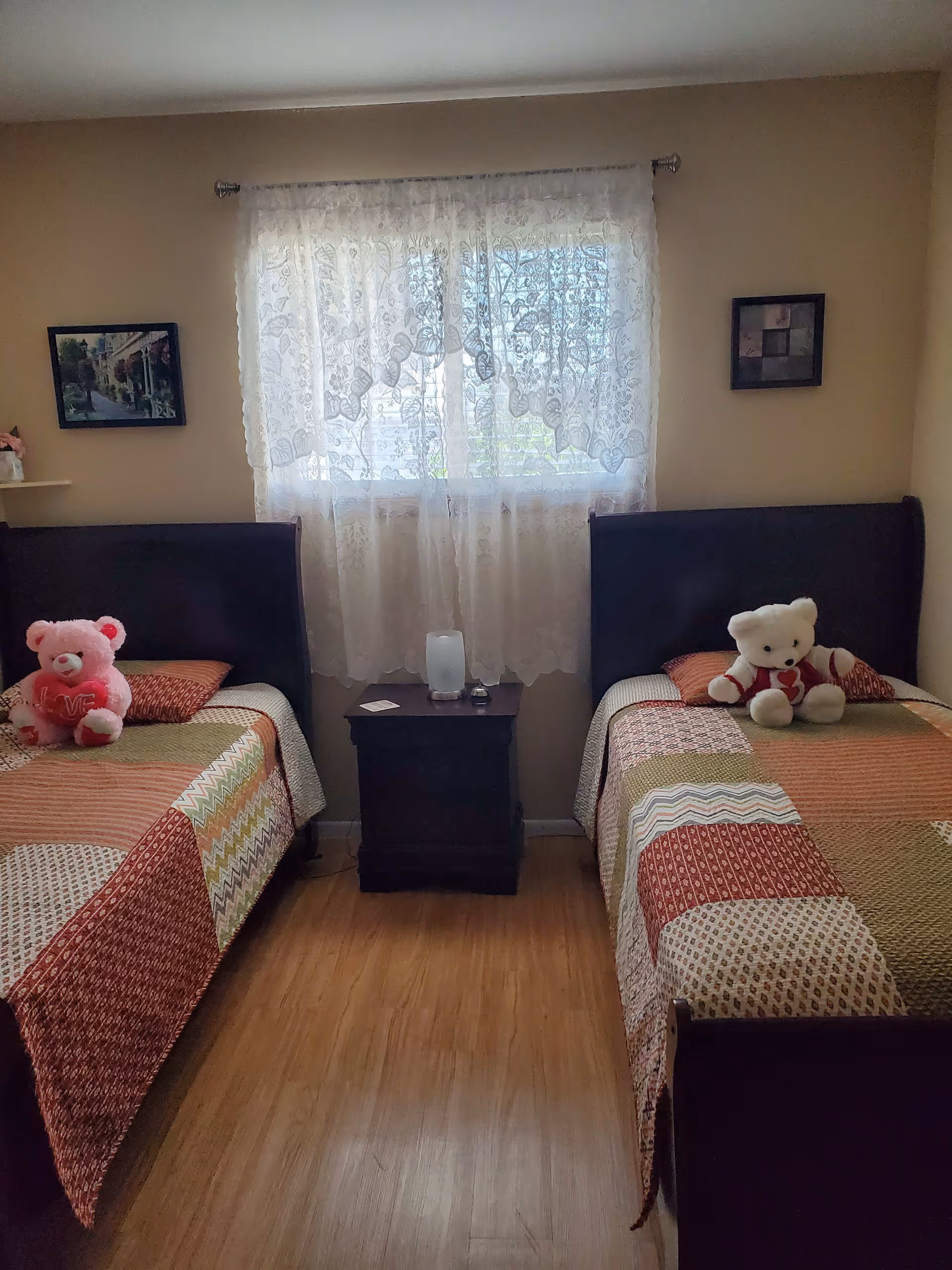 Small bedroom with two twin beds, a nightstand between them under a lace-curtained window and stuffed teddy bears on each bed.