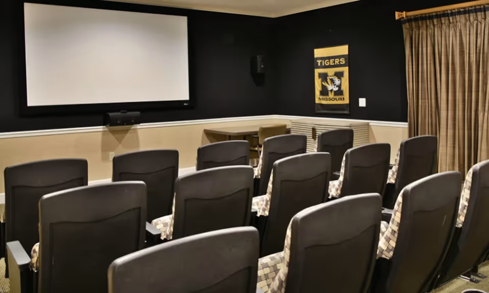 A small theater room with multiple rows of cushioned chairs facing a large white projection screen mounted on a black wall. There is a Tigers Missouri banner on the right wall and a window with closed curtains beside it.