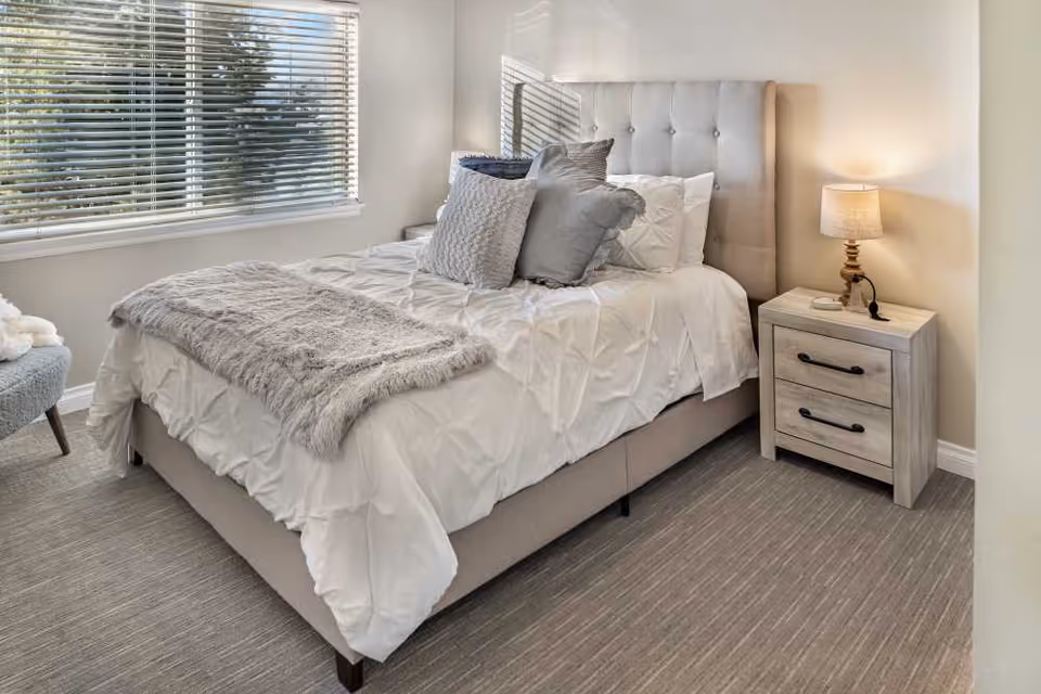Well-lit bedroom with a tufted headboard bed dressed in white linens, gray throw and pillows, a nightstand with lamp, and a window with blinds.