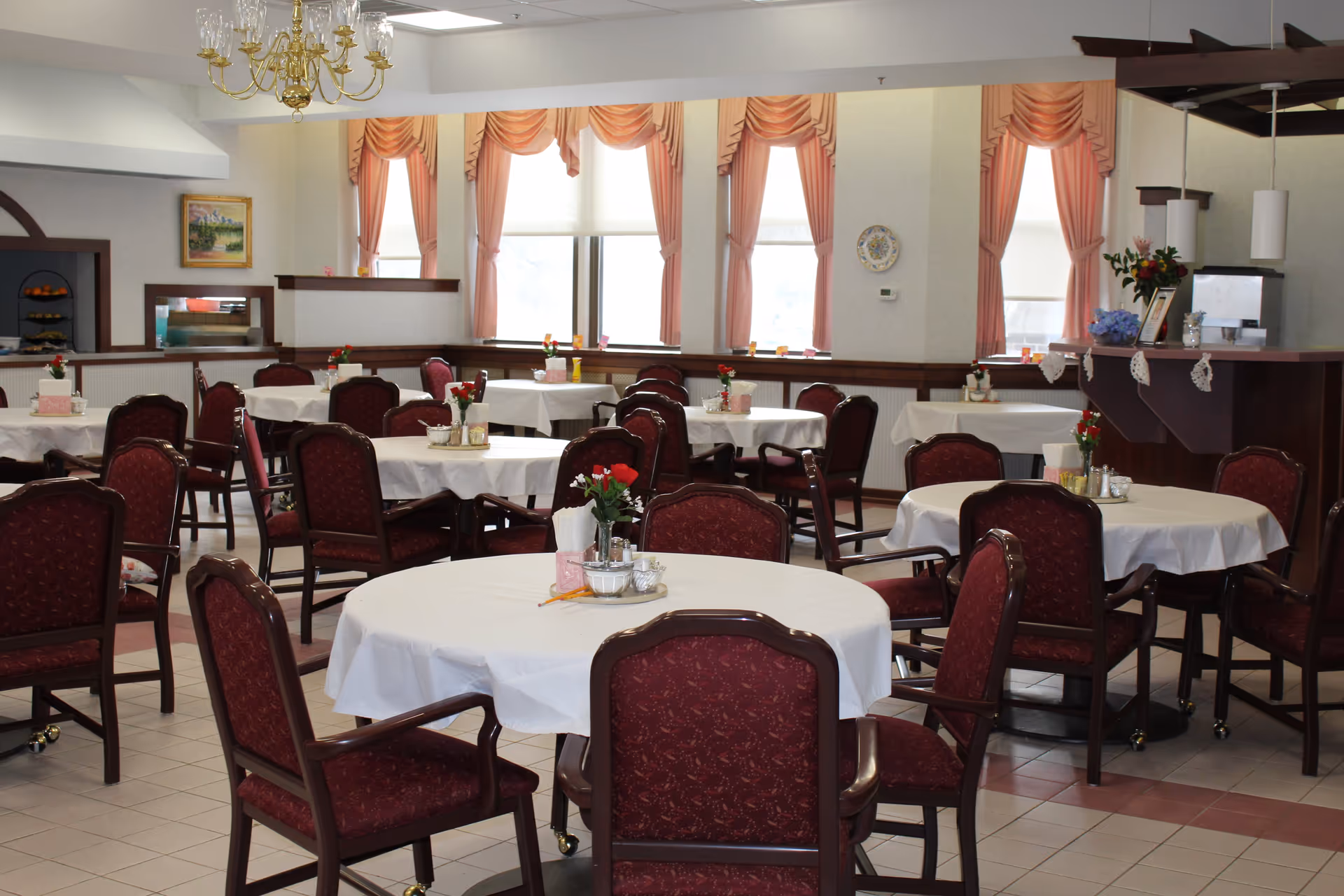 Bright dining room with round tables draped in white tablecloths, maroon upholstered chairs, floral centerpieces, and pink window drapes.