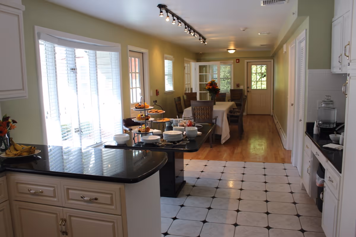 Interior view of a dining area and kitchen in an assisted living facility. The kitchen has white cabinets with black countertops and a tiled floor. A counter holds plates, bowls, and a tiered tray with snacks. The dining area features a table with chairs and a flower centerpiece, with windows and a door letting in natural light.