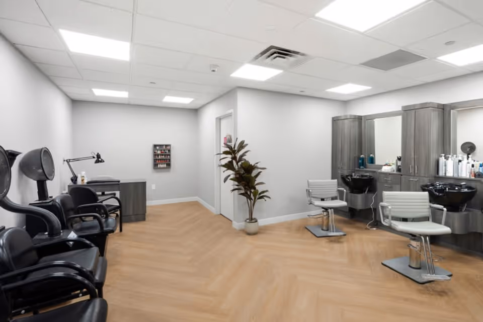 Interior view of a hair salon area in a senior living facility with two styling chairs in front of sinks and mirrors, black salon chairs with hair dryers on the left, a small desk with a lamp, and a plant in the corner. The room has light gray walls and wood-patterned flooring.