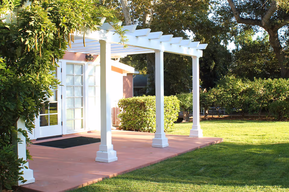 Sunlit patio with a white pergola in front of glass-paneled doors and a manicured lawn.