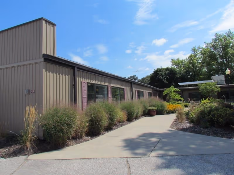 Exterior view of a single-story building with beige siding and purple window shutters, surrounded by landscaped bushes and plants along a concrete walkway under a partly cloudy blue sky.