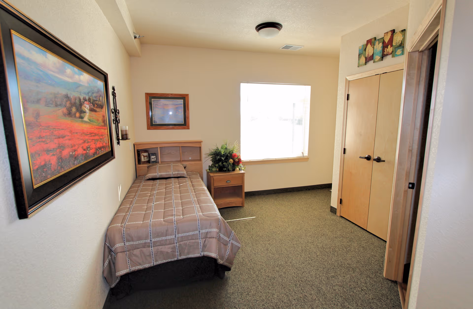 A small, simple bedroom with a single bed covered in a brown checkered bedspread, a wooden nightstand with a floral arrangement, a window letting in natural light, a closet with double wooden doors, and framed artwork on the walls.