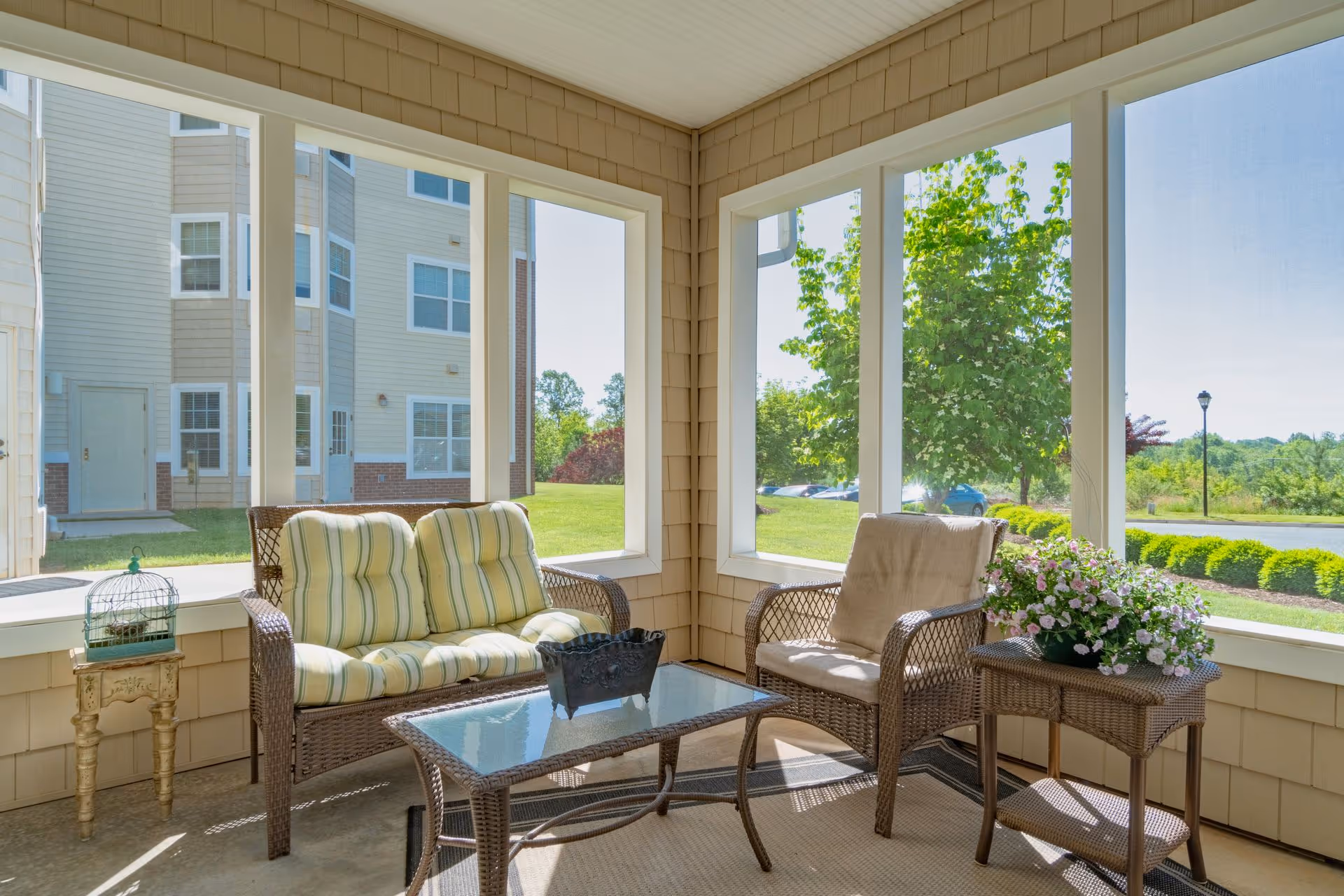 A bright and cozy screened-in porch area with wicker furniture including a cushioned loveseat and chair, a glass-top coffee table, a small side table with a potted flowering plant, and a decorative birdcage on a small wooden stand. Large windows provide views of a green lawn, trees, and a building exterior.