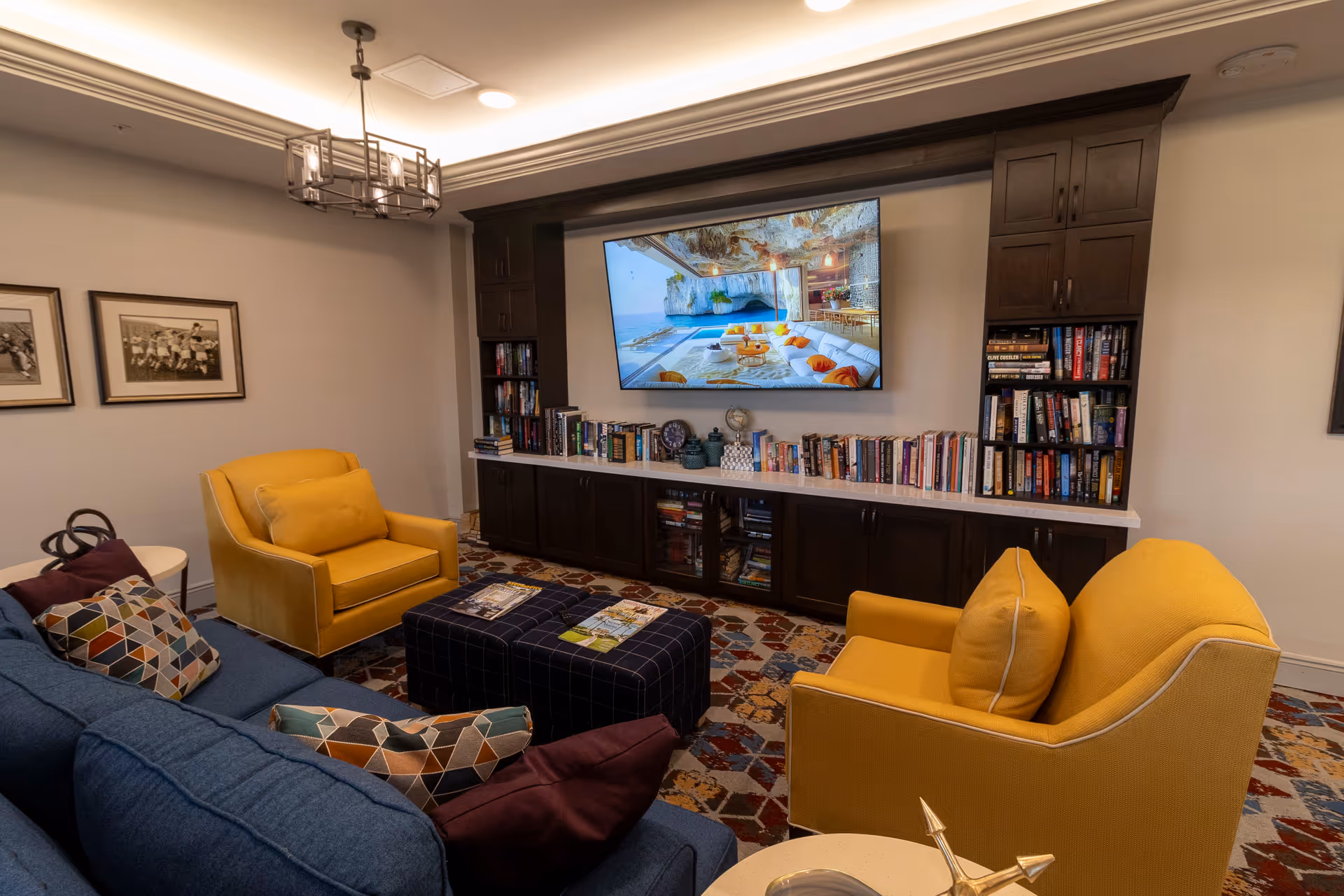 Cozy communal living room with a blue sofa, two yellow armchairs, patterned ottoman, built-in bookshelves and a wall-mounted TV.