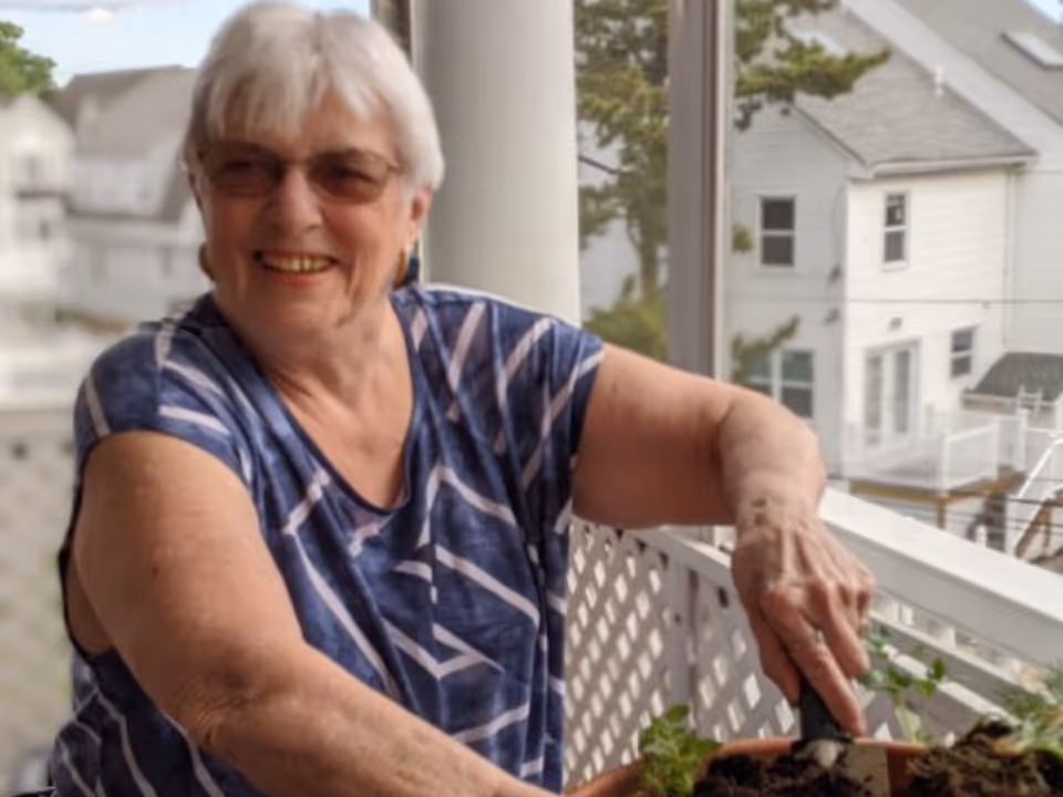 An elderly woman with short white hair and glasses is smiling while tending to a potted plant on a balcony with white lattice railing. Behind her, there are residential houses and trees visible through the balcony's glass enclosure.
