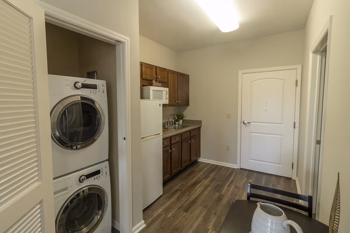 Interior view of a small kitchen area with wooden cabinets, a white refrigerator, a microwave, and a sink. To the left, there is a stacked washer and dryer unit inside a closet. The floor is wood-style laminate, and there is a white door at the end of the room. A small table with a white pitcher is partially visible in the foreground.