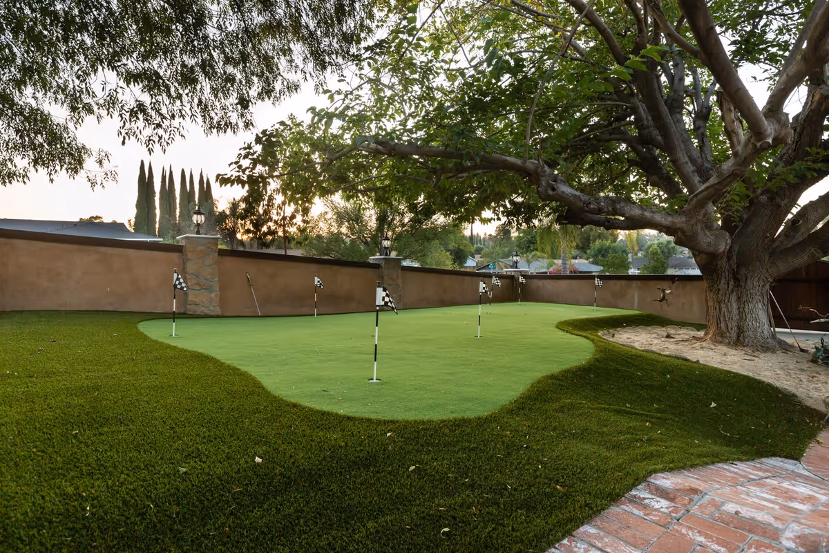 A backyard putting green with several small checkered flags on poles, surrounded by artificial grass and a large tree on the right side. A brick pathway is visible in the foreground, and a stone wall with lanterns lines the background.