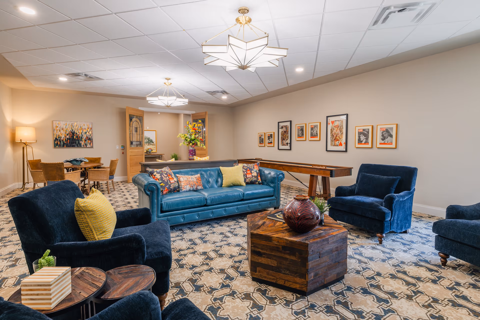 A spacious and well-lit living room area featuring a blue leather sofa with colorful cushions, three dark blue armchairs with yellow pillows, a wooden coffee table with a decorative vase, and a patterned carpet. The room has beige walls adorned with framed pictures and a shuffleboard table along one wall. In the background, there is a small dining table with four chairs and a floor lamp, with double wooden doors leading to another room.