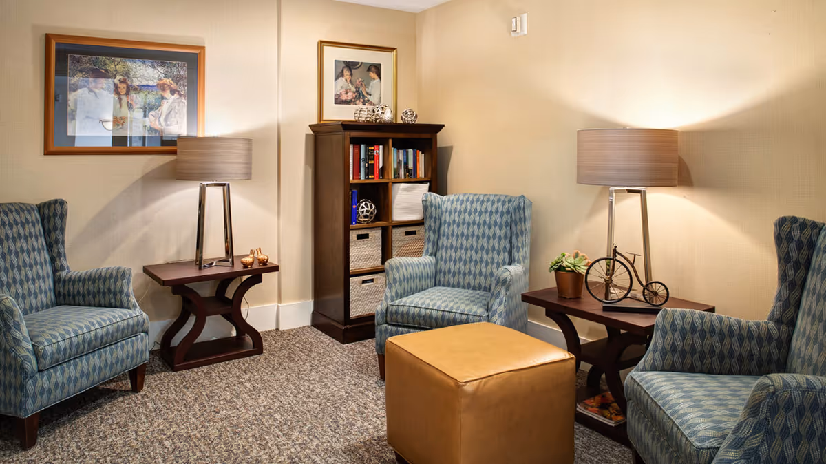 A cozy sitting area with three patterned blue armchairs arranged around a tan leather ottoman. There are two dark wooden side tables with modern lamps, a small decorative bicycle, and a potted plant. A wooden bookshelf with books and baskets is against the wall, which is decorated with two framed paintings.