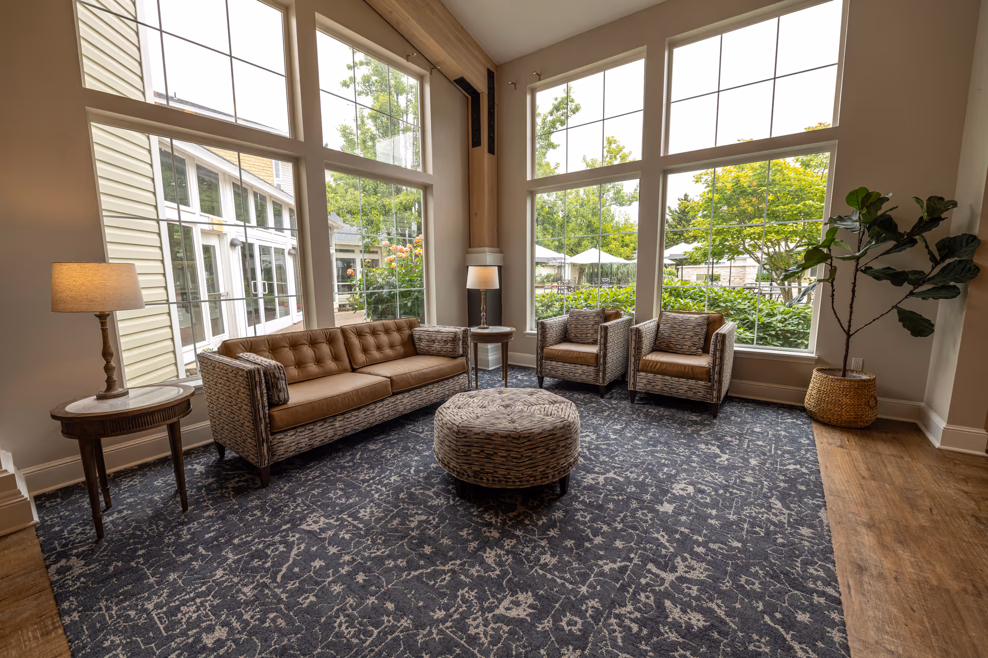 Sunlit seating area with a wicker sofa, two armchairs, a round ottoman, lamps, large windows, and a potted plant overlooking a garden.