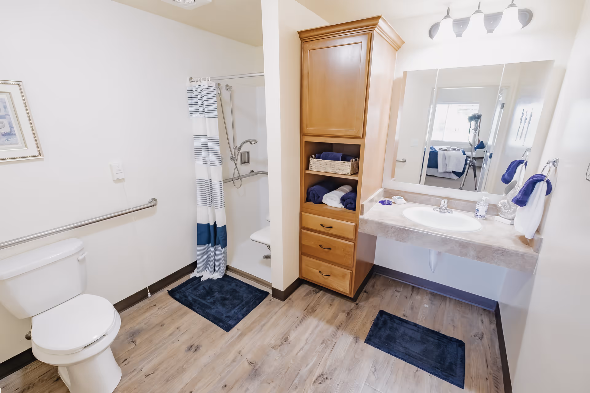 A clean and bright bathroom with a toilet, a walk-in shower with a striped shower curtain, a wooden cabinet with shelves and drawers holding folded towels, and a sink with a large mirror above it. The floor has a wood-like finish with two dark blue bath mats.