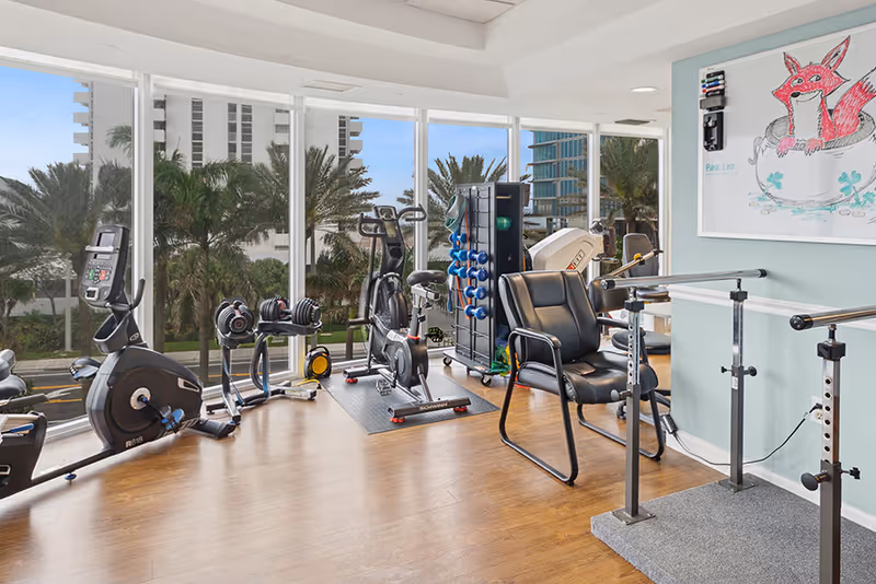 A bright fitness room with large floor-to-ceiling windows showing palm trees and buildings outside. The room contains exercise equipment including stationary bikes, dumbbells, and a rack of small weights. There is also a black chair and parallel bars for physical therapy or balance exercises. A whiteboard with a drawing of a red fox in a teacup is mounted on the light blue wall.