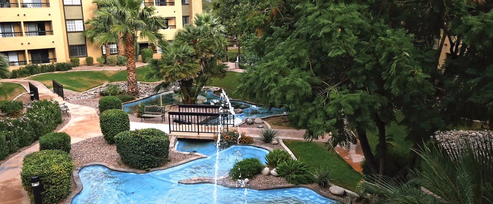 A landscaped outdoor area in a senior living facility featuring a winding blue water feature with a small fountain, surrounded by green bushes, palm trees, and other plants. There are paved walkways, benches, and a multi-story building with balconies in the background.
