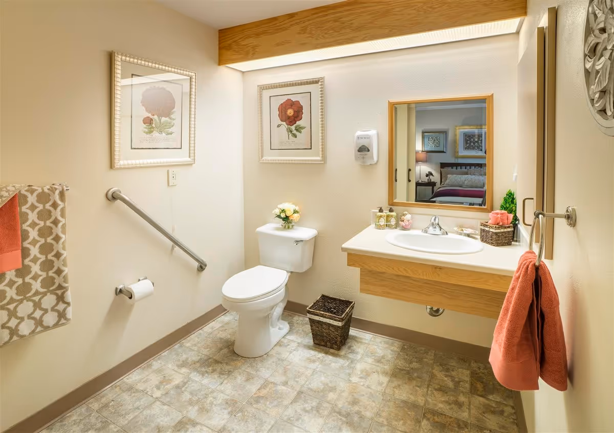 A clean and well-lit bathroom with a white toilet, a sink with a wooden cabinet, a mirror above the sink, and two framed floral pictures on the wall. There are coral and patterned towels hanging on the walls, a small basket on the floor, and a soap dispenser mounted on the wall.