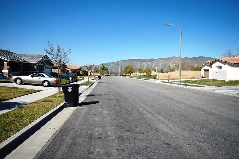 A wide residential street with houses on both sides, a few parked cars, trash bins along the sidewalk, and mountains visible in the background under a clear blue sky.