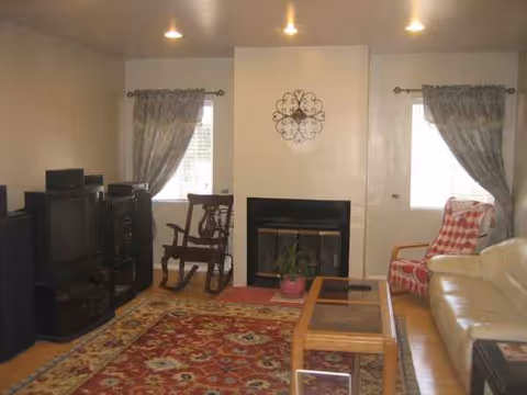 A cozy living room with cream sofa, coffee table, rug, fireplace, rocking chair, and draped windows.
