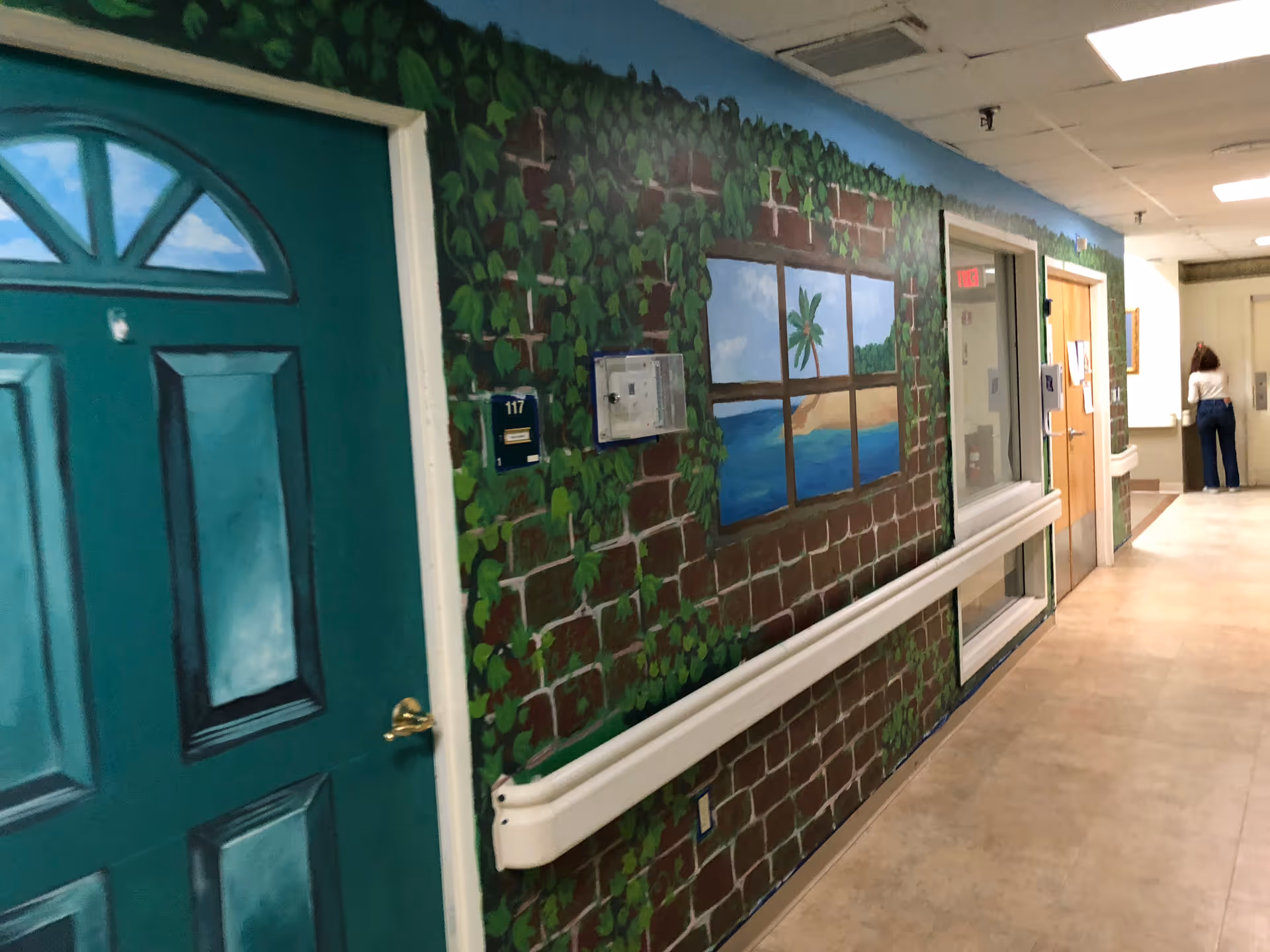 A hallway in a senior living facility with a green door labeled 117 on the left. The wall is painted with a mural depicting a brick wall covered with green ivy and a window showing a tropical beach scene with a palm tree and ocean. There is a handrail along the wall and a person standing at the far end of the hallway near a counter or desk.