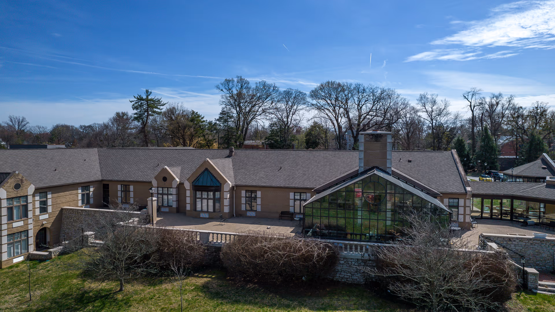 Exterior view of a single-story building with a large glass section and a chimney, surrounded by leafless trees and bushes under a clear blue sky.