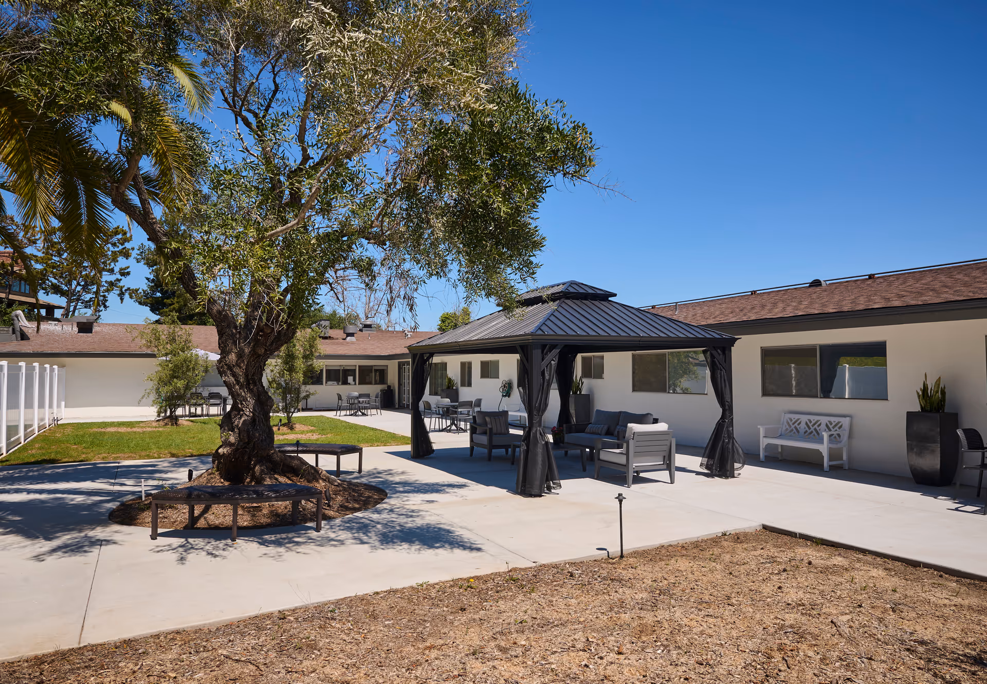 Outdoor courtyard with a central tree, circular bench, a gazebo with patio seating, and single-story building facades.