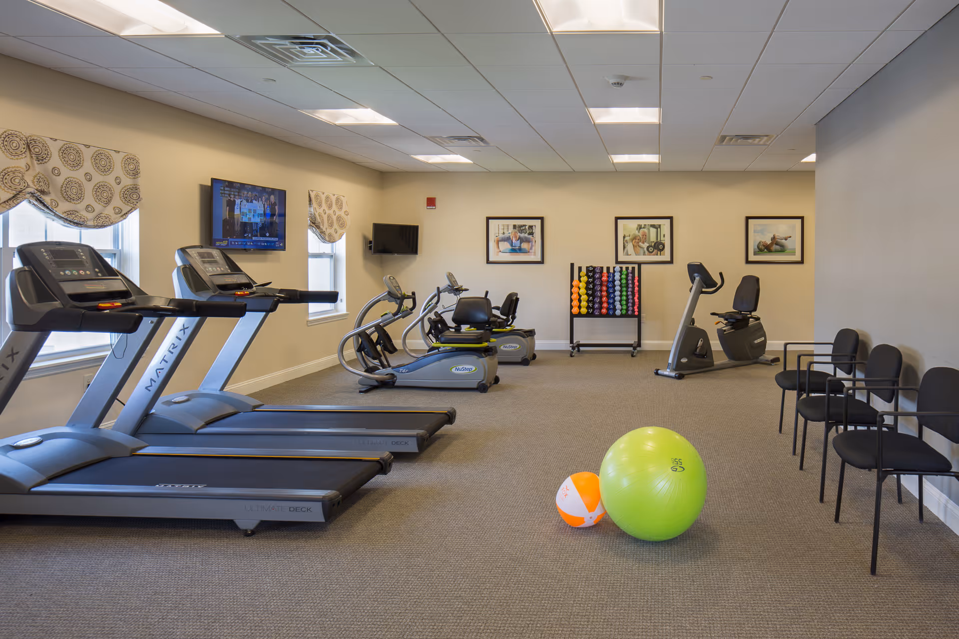 A senior living facility exercise room with two treadmills, two recumbent exercise bikes, a rack of colorful dumbbells, a green exercise ball, and an orange and white medicine ball. There are four black chairs lined up against the right wall and two windows with patterned valances on the left wall. A flat-screen TV is mounted on the wall between the windows, and three framed pictures hang on the far wall.