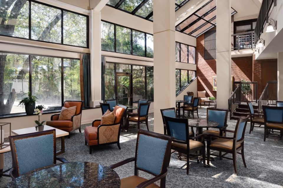 A bright and spacious common area in Woodland Heights Senior Living with large floor-to-ceiling windows allowing natural light to fill the room. The space features multiple seating arrangements including blue cushioned chairs around small round tables and orange cushioned armchairs with pillows near a window. There are plants on some tables and a staircase leading to an upper level. The walls include large windows and a brick accent wall.