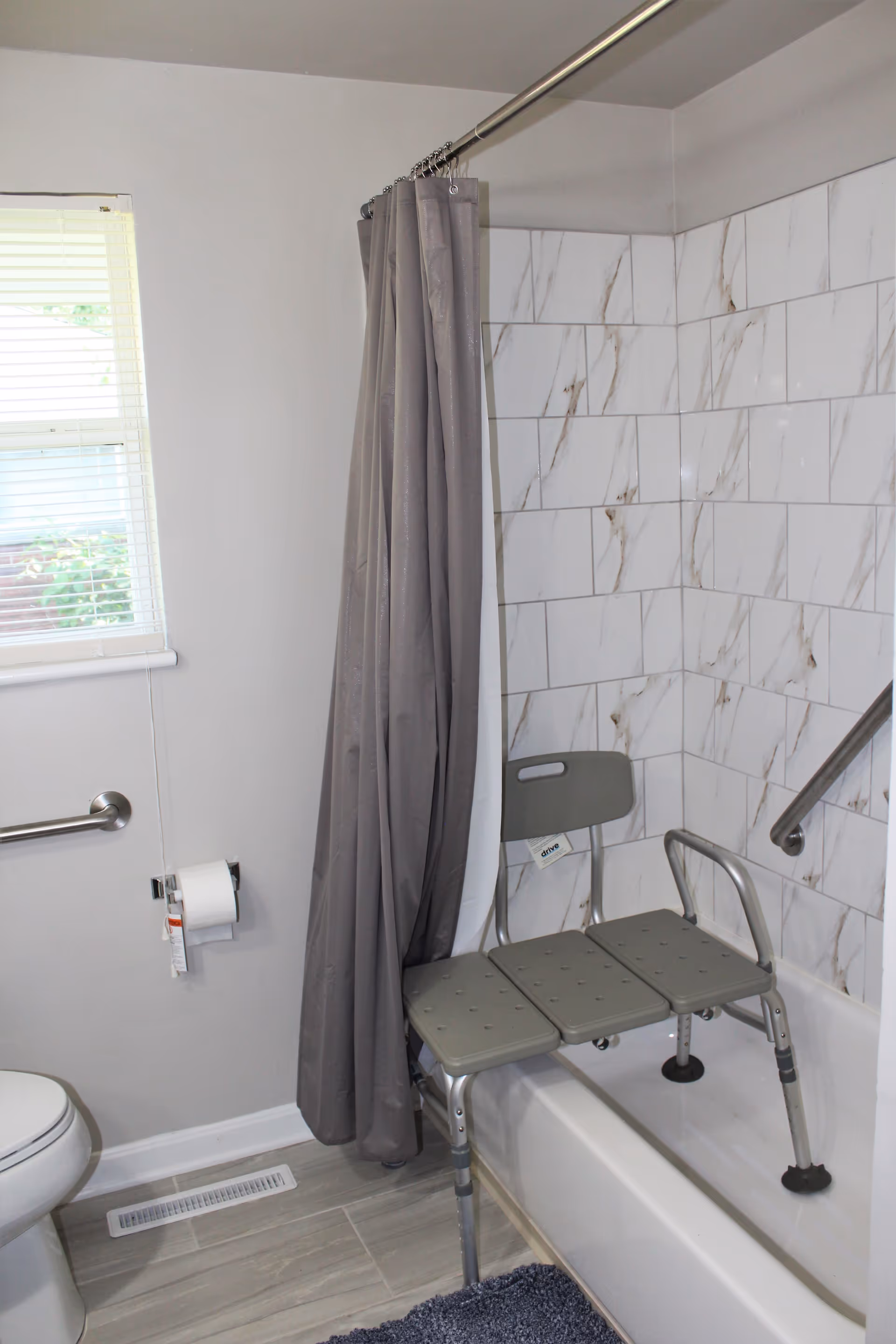A bathroom with a white toilet, a window with blinds, a gray shower curtain, and a bathtub equipped with a gray shower bench and grab bars. The bathtub walls are tiled with white tiles featuring subtle brown veining.