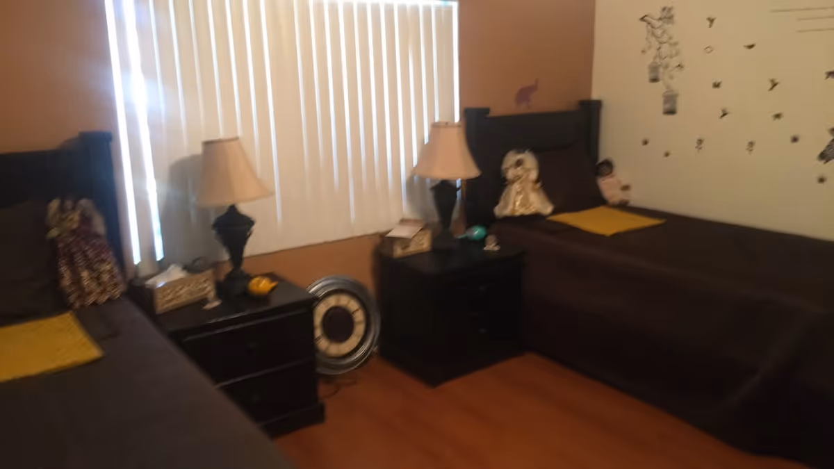 A bedroom with two single beds covered in dark brown bedding, each with a yellow blanket at the foot. Between the beds are two black nightstands, each with a lamp and decorative items. The room has vertical blinds covering the window and wooden flooring. There are dolls placed on the beds and wall decals of birds and flowers on one wall.