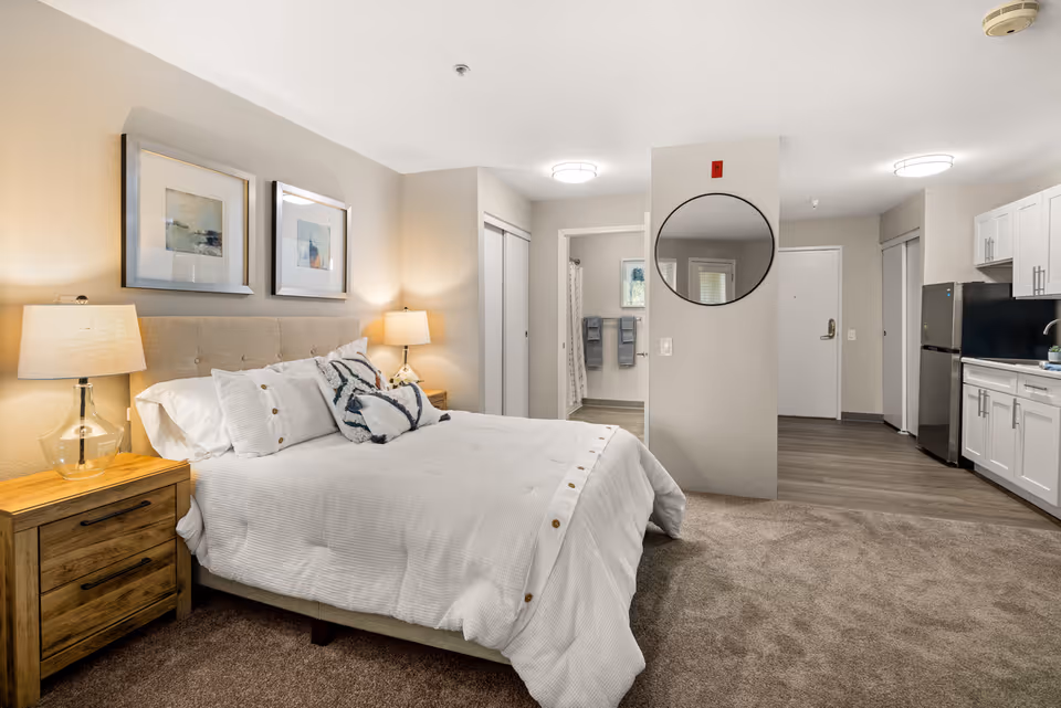 Furnished studio-style bedroom with a made bed, bedside tables and lamps, a kitchenette with refrigerator, and a bathroom visible through an open doorway.